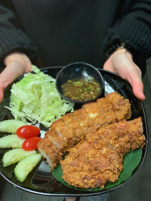 A server holding a plate of Crispy Pork Belly with lettuce, tomato, cucumber, and dipping sauce.